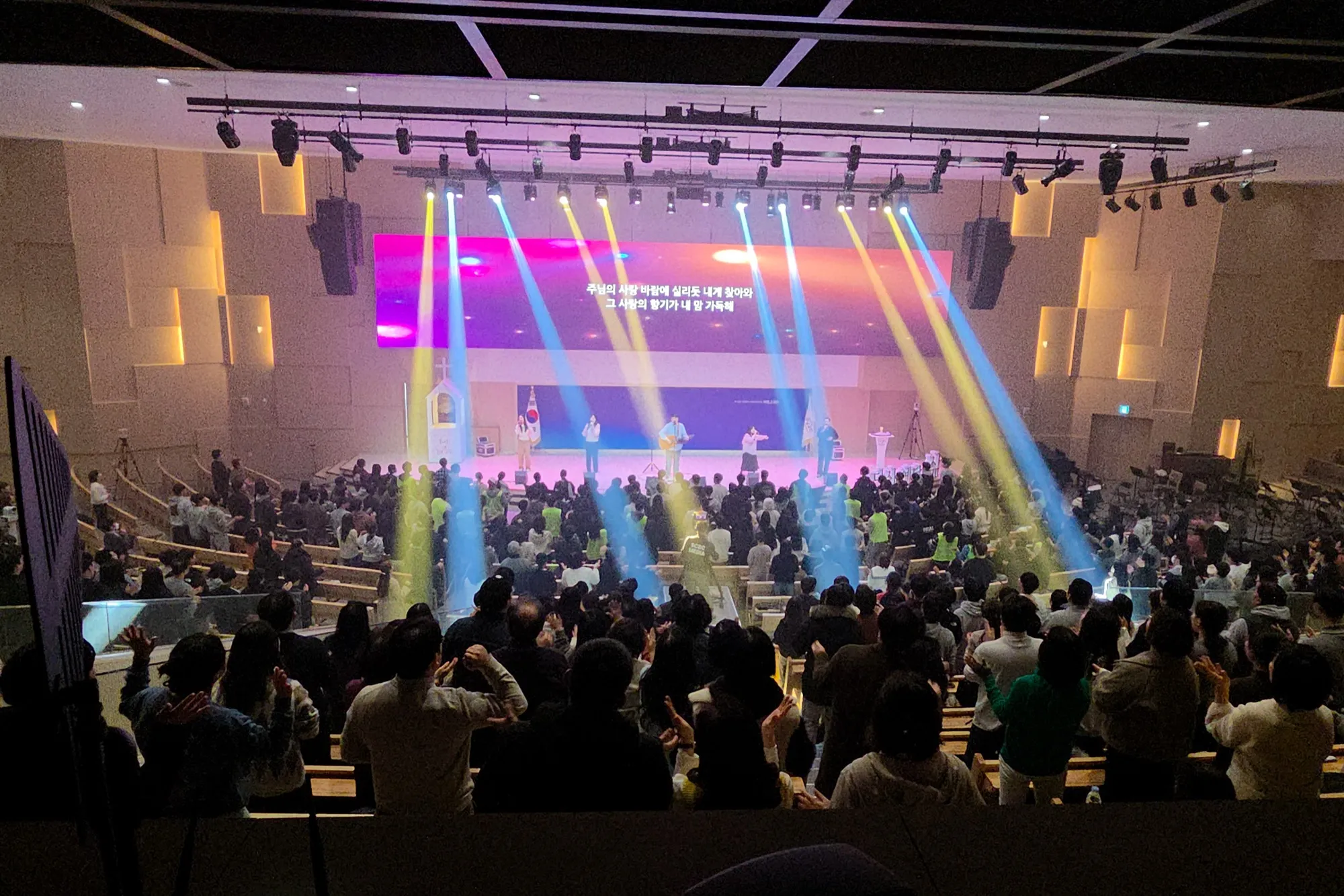 A large auditorium seen from the balcony. Markers Worship on stage under yellow and blue lights, the congregation clapping and singing