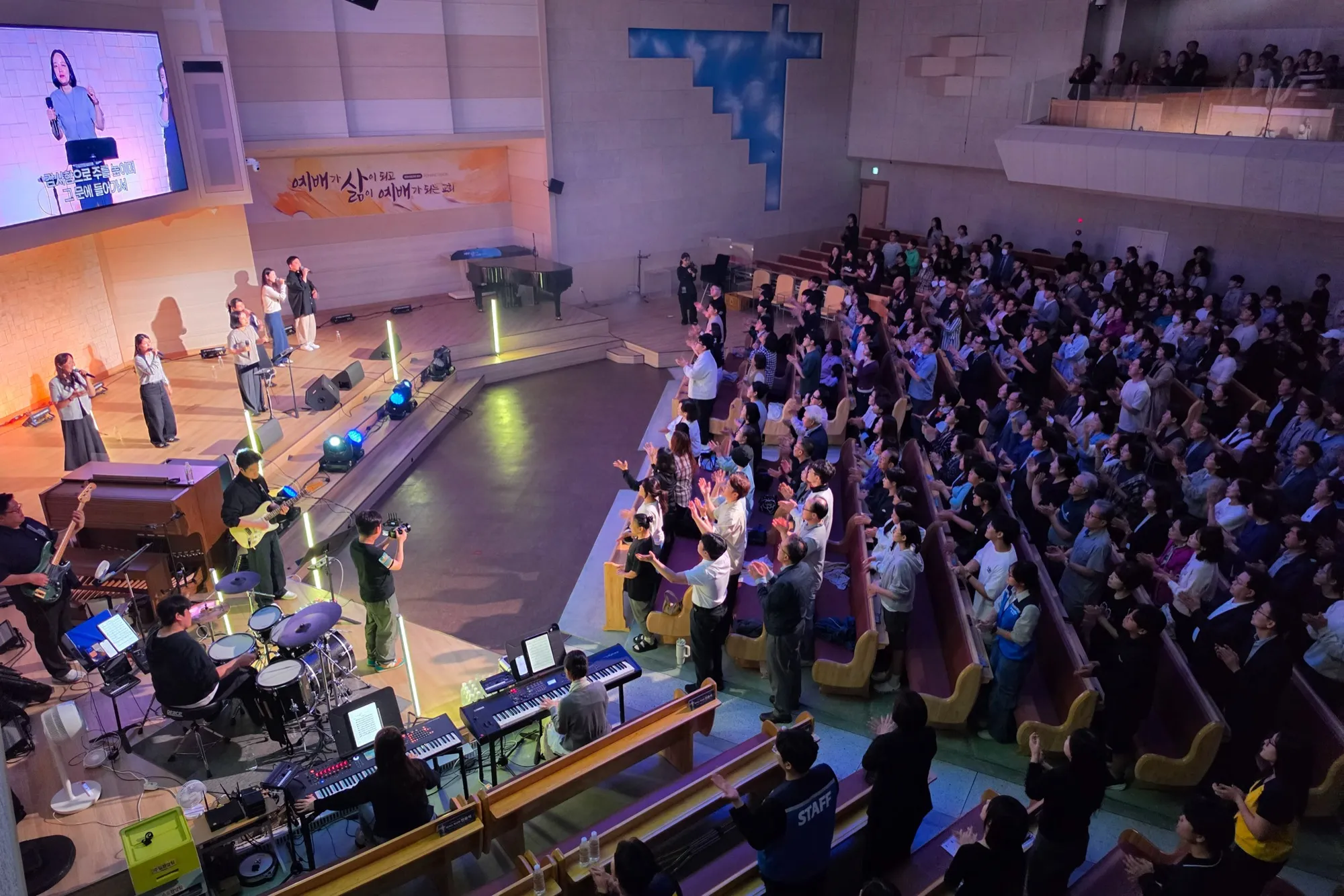A sanctuary seen from above. The band and vocalists lead worship as the congregation stands with hands raised
