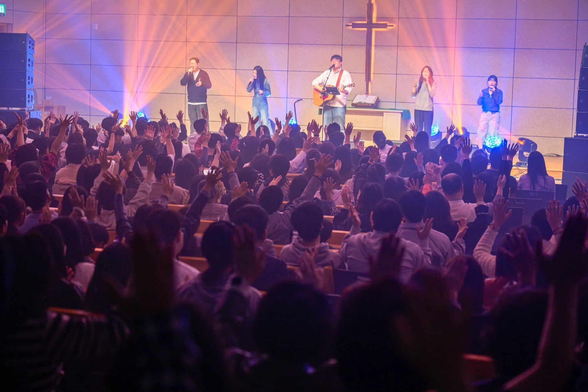 A worship leader with a guitar and vocalists singing in front of a cross, the congregation raising their hands