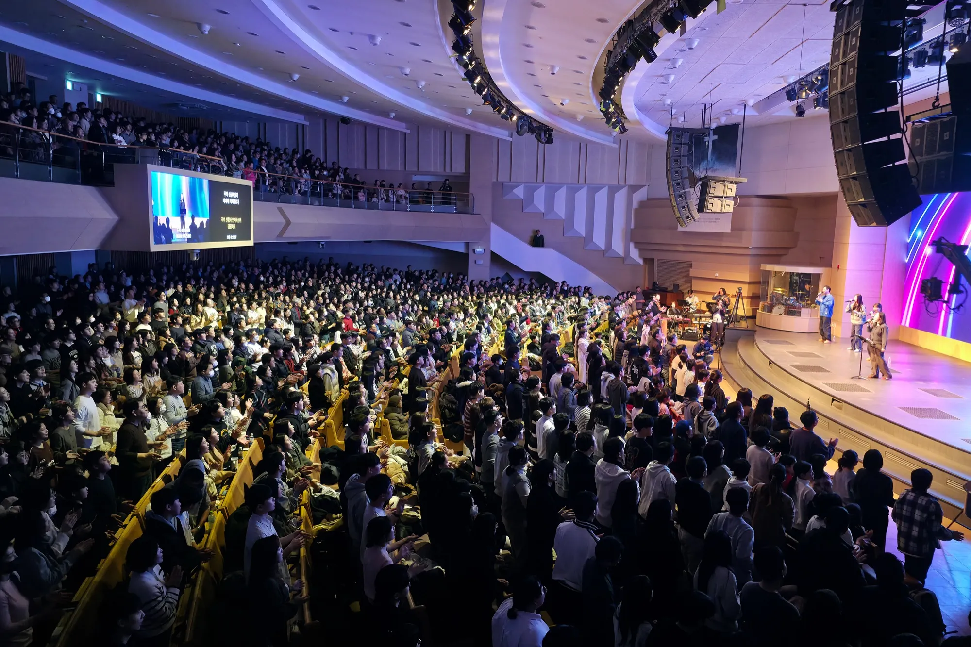A packed two-level auditorium, the congregation standing in worship with Markers Worship on stage to the right